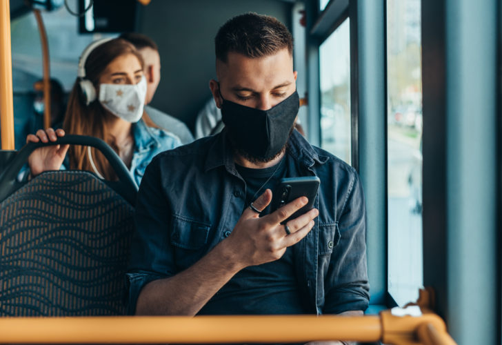 Handsome man wearing protective mask and using a smartphone while riding a bus and sitting on a distance from other passengers